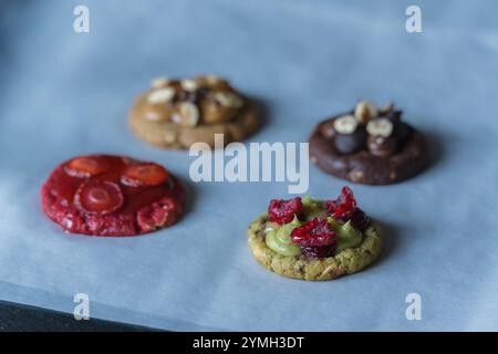 Hausgemachte runde Kekse mit verschiedenen Belägen. Himbeere, Erdbeere, Pistazien, Karamell, Schokolade, Erdnüsse, Erdnussbutter. Kekse auf Backpapier. Stockfoto