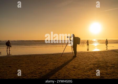 Fotograf in voller Silhouette steht am Stativ mit Kamera und langem Objektiv am Strand bei Sonnenuntergang. Eine ferne Figur im goldenen Licht fängt einen ruhigen Abend ein, bei dem Menschen entlang der Küste spazieren Stockfoto