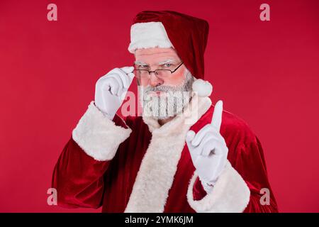 Porträt eines älteren Weihnachtsmanns, der die Brille anpasst, während er ein traditionelles rot-weißes Outfit vor einem leuchtend roten Hintergrund trägt und ein nachdenkliches und besinnliches Verhalten ausdrückt Stockfoto