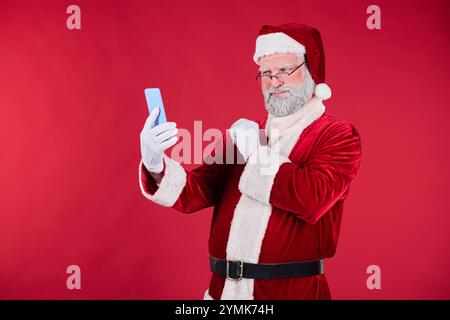 Der Weihnachtsmann lächelt, während er ein Selfie vor einem roten Hintergrund macht. Er trägt einen traditionellen roten Anzug, weiße Handschuhe und einen schwarzen Gürtel und wirkt fröhlich und festlich Stockfoto