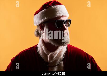 Mann mit weißem Bart, der Weihnachtsmann-Kostüm und Sonnenbrille trägt, die seitlich vor leuchtendem gelbem Hintergrund gerichtet ist, schafft festliche Feiertagsatmosphäre Stockfoto