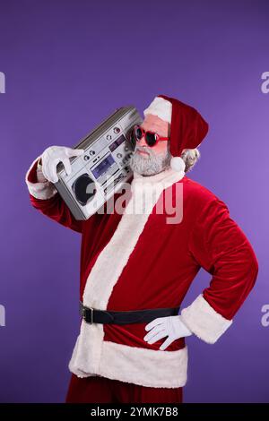 Der Weihnachtsmann trägt einen traditionellen roten Anzug und hält die Boombox auf der Schulter, während er eine Sonnenbrille trägt. Der Hintergrund ist schlicht violett und betont seine festliche Kleidung Stockfoto
