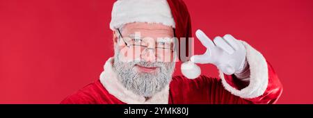 Porträt des Weihnachtsmanns, der direkt in die Kamera blickt, rot-weiß-Outfit mit weißen Handschuhen trägt, die Brille mit einer Hand justiert und vor rotem Hintergrund posiert Stockfoto