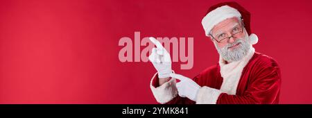 Älterer Mann, der als Weihnachtsmann gekleidet ist und vor rotem Hintergrund lächelnd und nach oben zeigt, während er traditionelle Tracht mit weißen Handschuhen und rotem Hut trägt Stockfoto