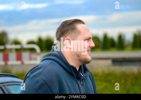 Ein Mann in einem dunklen Hoodie steht im Profil, lächelt sanft, mit einer ordentlichen Frisur und leichten Bartstoppeln. Stockfoto