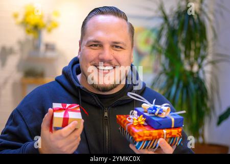 Ein fröhliches Porträt eines Mannes drinnen, der einen Stapel hell gewickelter Geschenke in der einen Hand und eine kleine Geschenkbox mit einem roten Band in der anderen hält. Stockfoto