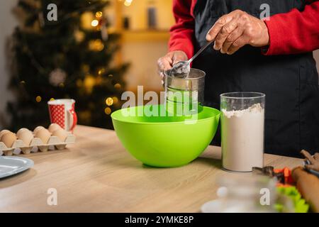 Hände eines älteren Mannes in Schürze, der Mehl für hausgemachte Backwaren über grüner Schüssel siebt, während er am Heiligabend am Küchentisch steht Stockfoto