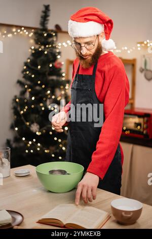 Junger Mann mit Weihnachtsmütze und Schürze, der Teig für weihnachtskuchenkekse und Weihnachtsbonbons macht, bärtiger Kerl, der während der Winterferien kocht Stockfoto