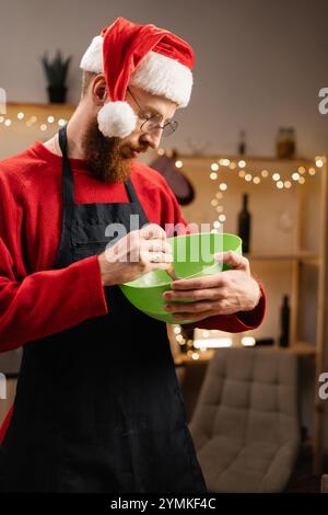 Mann mit Weihnachtsmann-Hut, der Teig in grüner Schüssel mischt, sich auf das Weihnachtsbacken in der festlichen Küche vorbereitet, Lichter funkeln herum. Fokussiert auf das Machen Stockfoto