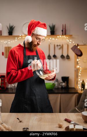 Baker in Santa hat Eier in einer Glasschale verquirlt und Zutaten für das Backen in seiner Küche zubereitet. Konzept des Hausbacken und der kulinarischen Zubereitung. Stockfoto