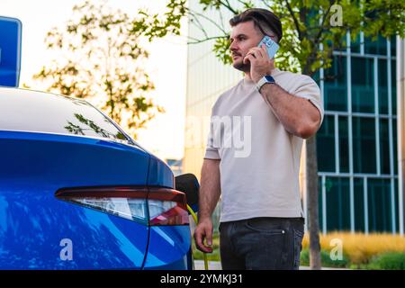 Der Mann steht neben seinem Elektroauto und spricht über sein Handy, während es in der Stadt auflädt Stockfoto