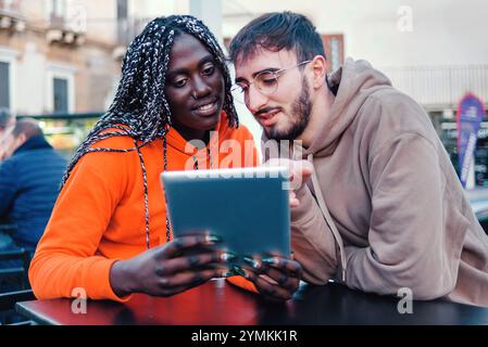 Multirassische Freunde teilen digitale Inhalte auf einem Tablet-Computer, während sie in einem Café im Freien sitzen. Afrikanerin in orangem Hoodie und kaukasischer Mann Collabo Stockfoto