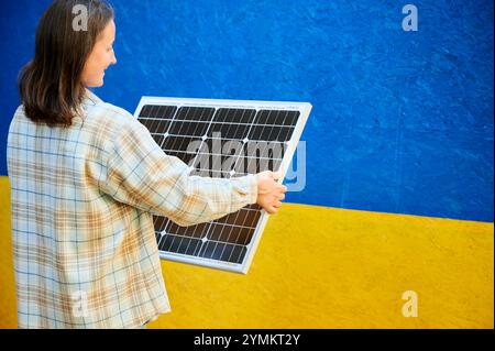 Frau, die Solarpanel zum Laden hält. Die lächelnde Frau trägt ein kariertes Hemd über weißem Oberteil und steht vor leuchtend blauem und gelbem Hintergrund der ukrainischen Flagge. Stockfoto