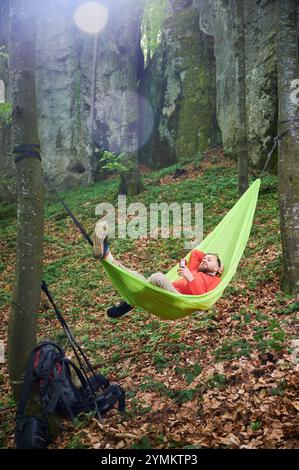 Person, die sich in einer hellgrünen Hängematte zwischen zwei Bäumen in einem üppigen, grünen Wald in der Nähe von Felsen entspannt. Man Tourist hält einen Drink und genießt einen friedlichen Moment inmitten der Natur. Rucksack und Zahnradauflage in der Nähe. Stockfoto