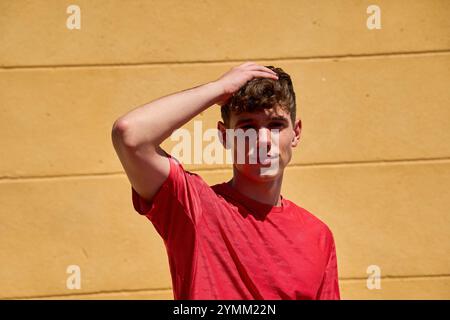 Ein junger Mann in einem roten Hemd steht vor einer gelben Wand. Er schaut hoch und berührt sein Haar. Das Konzept von Gelassenheit und Selbstvertrauen Stockfoto