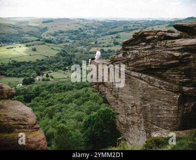 Ein Mann sitzt auf einem Felsen am Curbar Edge mit Blick auf ein Tal. Die Szene ist friedlich und ruhig, der Mann genießt die Aussicht und die Ruhe des Ar Stockfoto