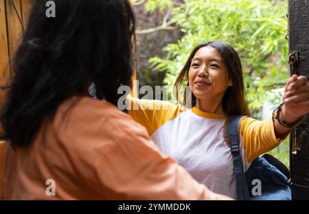 asiatische Teenagerin grüßt Großmutter mit Umarmung zu Hause, lächelt und hält Rucksack. Familie, Homecoming, generationenübergreifend, warm, Zuneigung, Konz Stockfoto