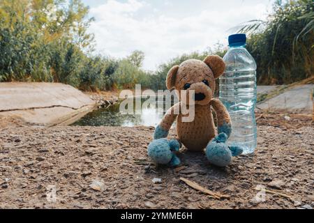 Ein Teddybär und eine Flasche Wasser stehen an einem bewölkten Sommertag neben einem trocknenden Bewässerungskanal. Konzeptionelle Darstellung des Klimawandels Stockfoto