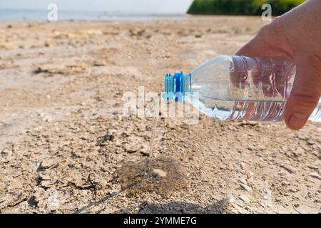 Plastikflasche in der Hand Nahaufnahme. Wasser fließt aus der Flasche auf die trockene Oberfläche des Bodens eines ausgetrockneten Sees. Konzept des Klimawandels Stockfoto
