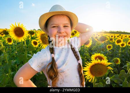 Ein fröhlich lächelndes Mädchen steht auf einem Feld mit Sonnenblumen und hält ihren Hut mit der Hand. Sonnenuntergang oder Sonnenaufgang im Sommer Stockfoto