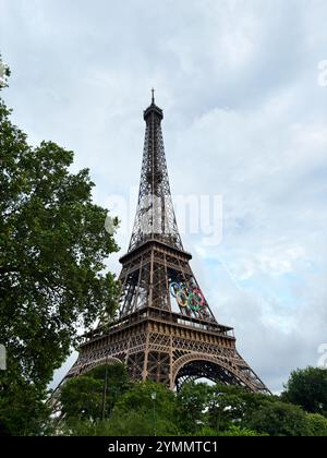 Paris, Frankreich - 19. Juni 2024: Der berühmte Eiffelturm ist mit olympischen Ringen geschmückt, vor dem Hintergrund eines bewölkten Himmels und grüner Bäume. Ein Symbol von Stockfoto