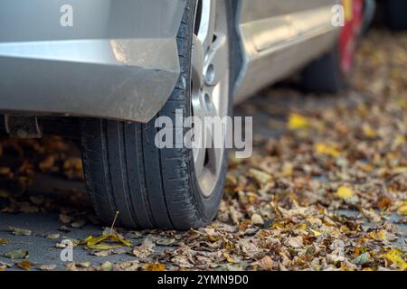 Platter Reifen auf einem Bett aus bunten Herbstlaub in der Nähe eines geparkten silbernen Autos Stockfoto