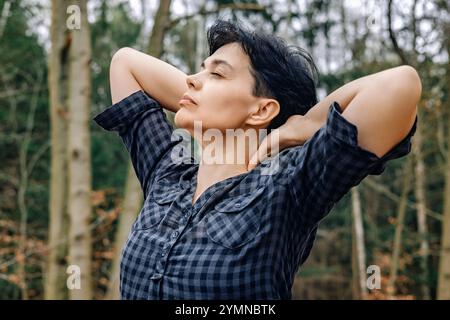 Glückliche Frau, 45-50 Jahre alt, trägt ein kariertes Hemd, genießt die frische Luft im Wald mit ihren Händen hinter ihrem Kopf Stockfoto