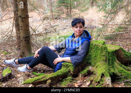 Eine Frau sitzt in einem Kiefernwald und lehnt sich auf einen Baumstamm. Der alte Stumpf ist mit grünem Moos bedeckt. Stockfoto
