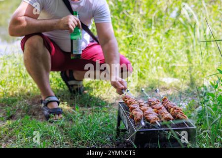 Ein Mann mit einer Flasche Bier grillt Schaschlik, während er vor einem Grill sitzt. Stockfoto