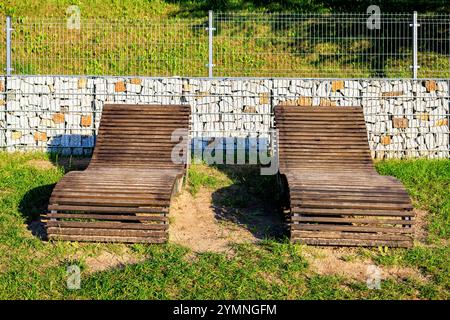 Zwei stationäre Sonnenliegen aus Holz auf dem Rasen eines Stadtstrandes. Vorderansicht vor einem Gobion-Hintergrund. Stockfoto