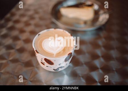 Schokoladentasse Cappuccino und Kuchen auf einem Tisch auf der Terrasse des Cafés draußen. Stimmungsvolle Kaffeezeit. Herzförmige Dessertdekoration Stockfoto