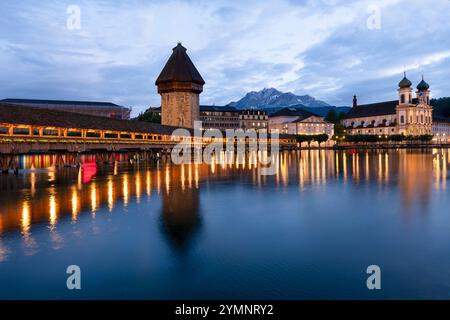 Beleuchtete überdachte Holzstege und Wasserturm, die sich im Wasser spiegeln, führen in der Abenddämmerung über den Fluss Stockfoto