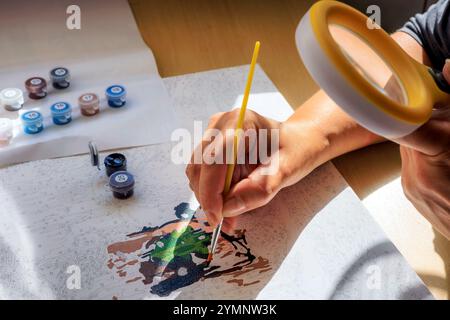 Malen nach Zahlen. Eine Frau malt ein Bild mit Zahlen mit einem Pinsel und schaut durch eine Lupe auf die Leinwand Stockfoto