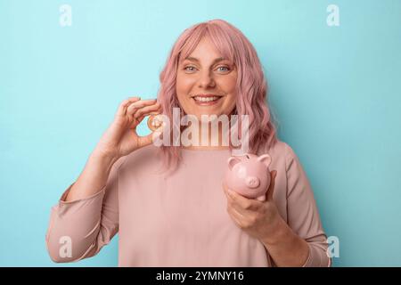 Junge kaukasische Frau, die Schweinebank und Bitcoin isoliert auf hellblauem Hintergrund hält und einlädt, mit der Hand zu kommen. Stockfoto