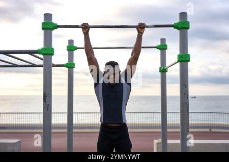 Junger schwarzer Mann, der bei Sonnenaufgang Calisthenics auf einer Pull-up-Bar am Meer vorführt Stockfoto