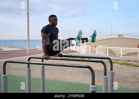 Junger schwarzer Mann, der Kalisthenics vorführt, sitzt auf parallelen Bars am Meer Stockfoto