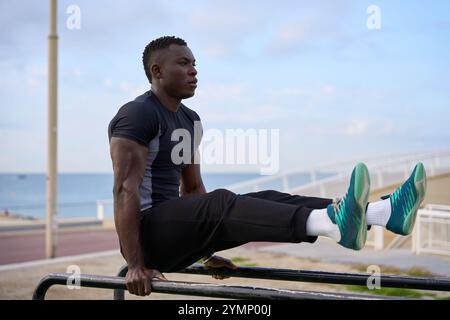 Junger schwarzer Mann, der Kalisthenics vorführt, sitzt auf parallelen Bars am Meer Stockfoto