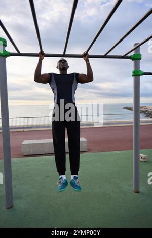 Junger schwarzer Mann, der Calisthenics-Pull-ups auf Outdoor-Fitnessgeräten am Meer durchführt Stockfoto