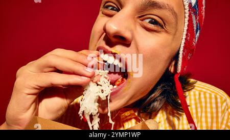 Nahaufnahme eines jungen Mannes im Winterhut emotional essende Wurst mit Sauerkraut auf rotem Hintergrund. Street Food im Winter. Schmutziges Gesicht im Senf Stockfoto