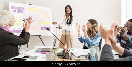 Ein enthusiastisches, vielfältiges Business-Team feiert den Erfolg während der Strategieplanung, wobei eine indische Moderatorin die Diskussion am Whiteboard leitet Stockfoto