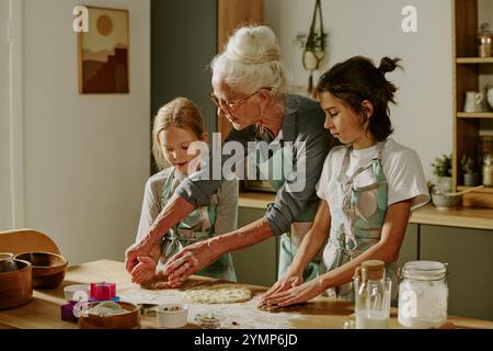 Seniorin, die zwei junge Mädchen in der Küche begleitet, während sie gemeinsam Teig zubereiten, konzentrierte sich auf das Backen von Keksen und genießen den Moment ohne Ablenkung Stockfoto