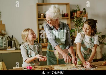 Ältere Frau, die mit zwei Kindern in der gemütlichen Küche backen und Spaß und lehrreiche Aktivitäten haben. Beide Kinder konzentrierten sich auf das Lernen und die Freude am Backen unter Anleitung der Senioren Stockfoto