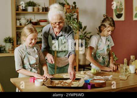 Ältere Frau mit zwei jungen Mädchen, die in der Küche backen, gemeinsam an Aktivitäten teilhaben und Momente teilen, in denen sie Kekse mit Teigschneidern auf Holztisch herstellen, die Freude darstellen Stockfoto