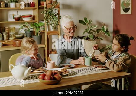 Ältere Frau, die schöne Zeit mit Enkelkindern am Frühstückstisch verbringt. Tee und warme Gespräche in gemütlicher Küche, umgeben von Pflanzen und Küchenutensilien Stockfoto