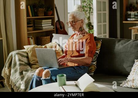 Ältere Frau mit weißem Haar und Brille, die auf dem Sofa sitzt und einen Laptop in einem gemütlichen Wohnzimmer benutzt und einen grünen Becher auf dem Tisch in ihrer Nähe aufgestellt hat Stockfoto
