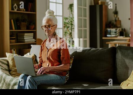 Ältere Frau mit weißem Haar, die auf einer Couch sitzt, einen Laptop in einem gut beleuchteten Wohnzimmer mit moderner Einrichtung benutzt und sich auf ihre Arbeit konzentriert Stockfoto