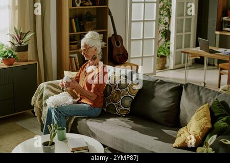 Eine ältere Frau strickt auf einem bequemen Sofa in einem gemütlichen, gut beleuchteten Wohnzimmer mit Pflanzen und Gitarre im Bücherregal und genießt einen ruhigen Moment Stockfoto