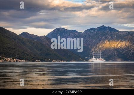 Ein ruhiger Blick auf die Bucht von Kotor in Montenegro, mit einem weißen Segelboot, das über ruhiges Wasser unter einem bewölkten Himmel gleitet, mit schroffen, sonnendurchfluteten mountai Stockfoto