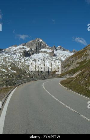 Gewundene Bergstraße in den Alpen mit schneebedeckten Gipfeln im Hintergrund unter hellblauem Himmel, ideal für Reise- und Naturthemen. Stockfoto