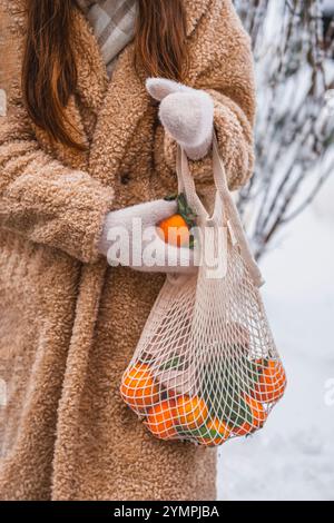 Hände einer jungen Frau, die im Winter eine Mesh-Einkaufstasche mit frischen Mandarinen hält. Keine Plastiktüte, kein Abfall, umweltfreundliches Konzept Stockfoto